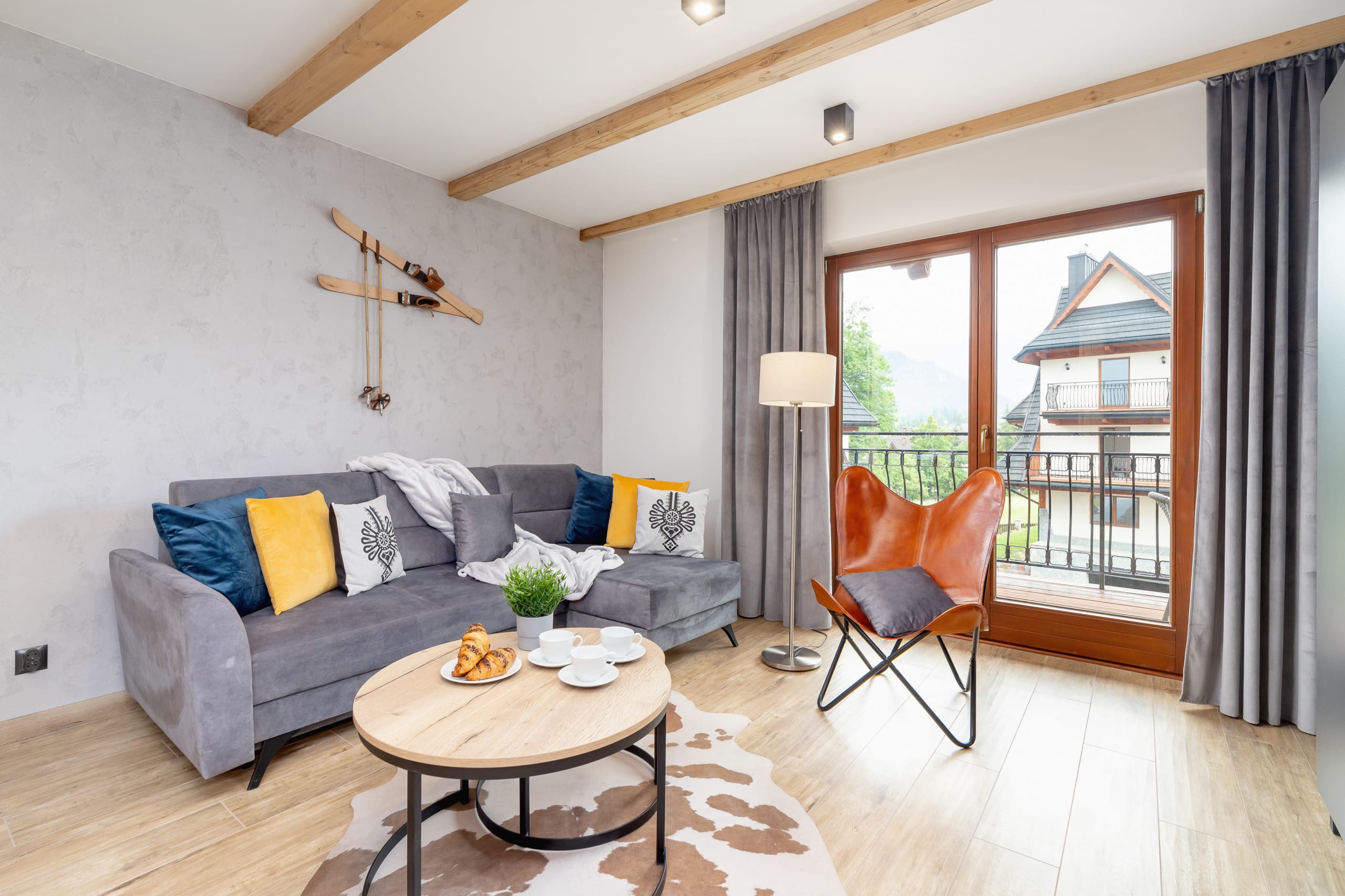 A modern living room with wooden beams on the ceiling, a sofa, armchairs, and a coffee table. Large windows provide natural light and views.