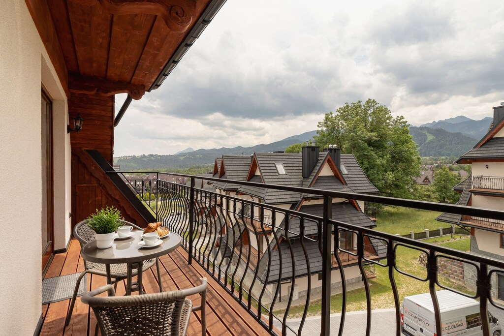 A wooden-floored balcony with a metal railing, overlooking a peaceful neighborhood with scenic mountain views.