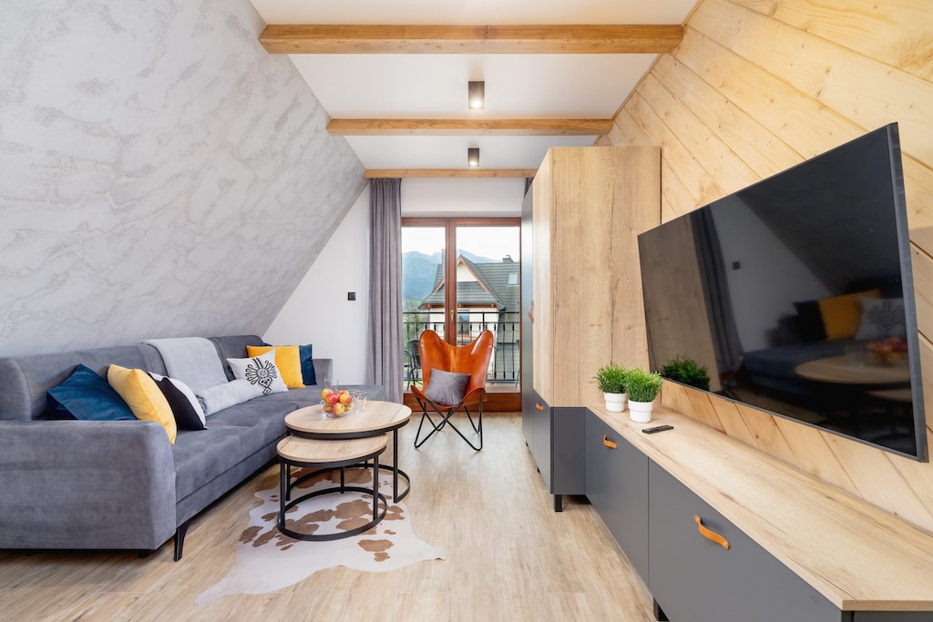 A cozy living room with a wooden dining table, chairs, and a sofa. Large windows let in natural light and offer a bright atmosphere.