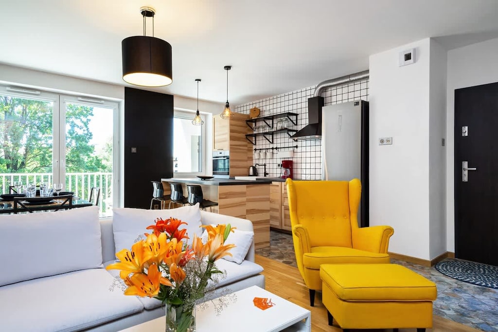 A stylish living room with a vibrant yellow armchair, a modern sofa, and a coffee table adorned with fresh flowers, surrounded by large windows that bring in natural light.