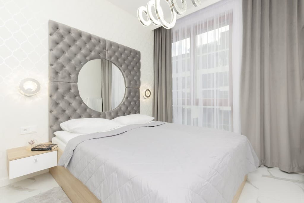 A bright bedroom with a plush upholstered headboard, grey bedding, and a round mirror above the nightstand, exuding a chic and cozy vibe.

