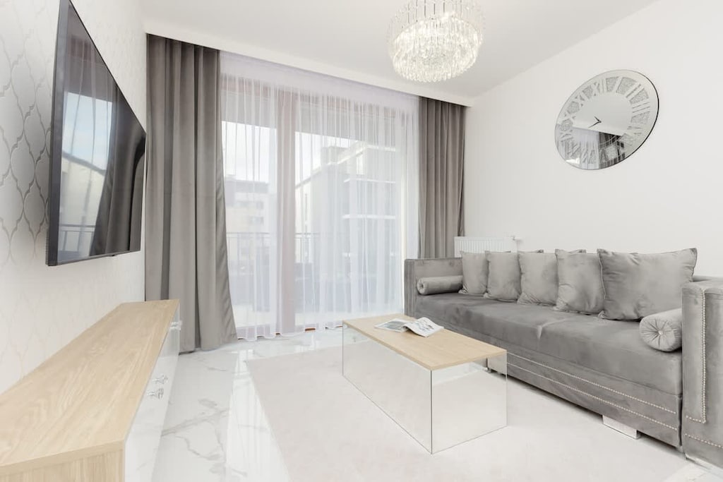 A spacious living room with a grey sofa, wall-mounted TV, and large windows covered with elegant curtains, providing a stylish and relaxing space.

