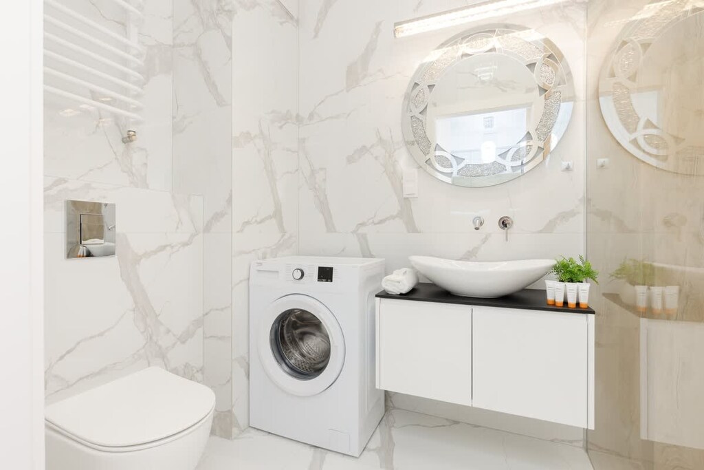A luxurious bathroom with marble tiles, a washing machine, a floating vanity, and a round illuminated mirror, showcasing a spa-like atmosphere.
