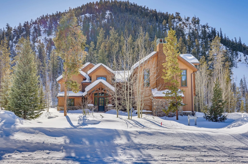 A wooden house with snow-covered roof and trees stands in front of a forested hill in winter.