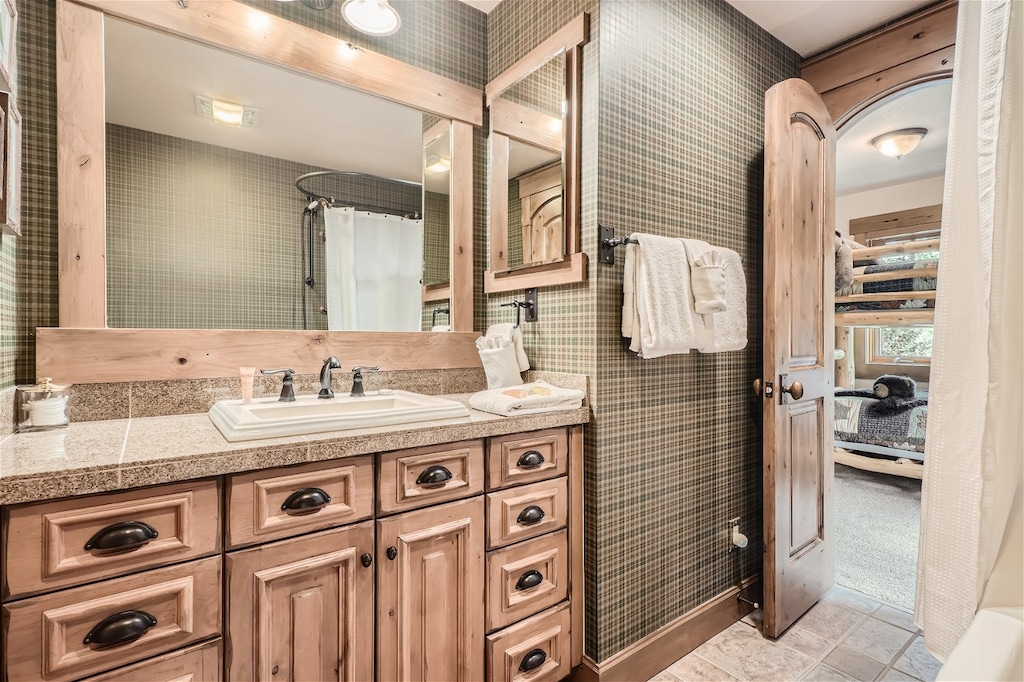 A rustic bathroom with wooden cabinets, granite countertop with a double sink, wall mirror, and a towel rack. A door leads to a room with bunk beds visible in the background.