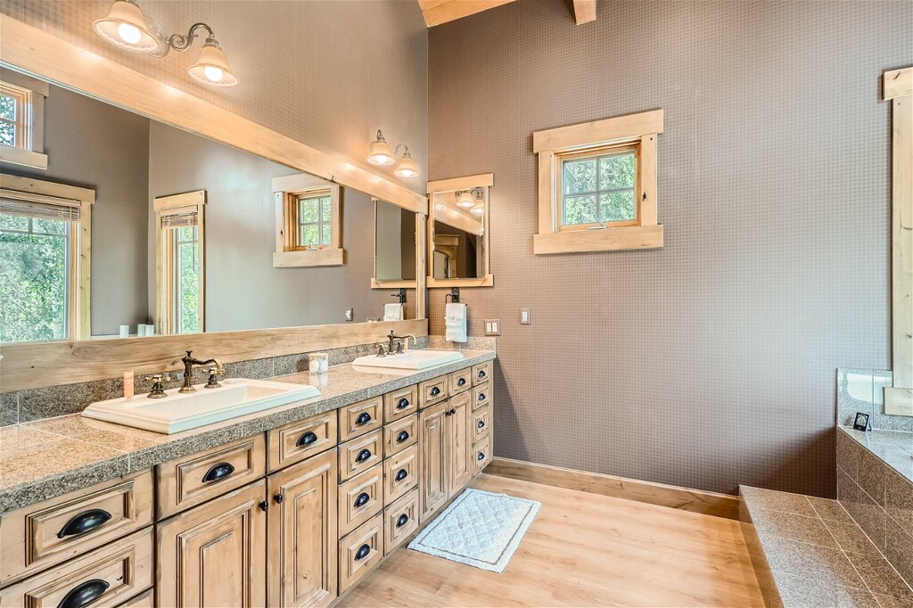 A spacious bathroom with a rustic design features double sinks, a large mirror, wooden cabinets, granite countertops, and a tiled bathtub. The room has wooden accents and windows offering natural light.