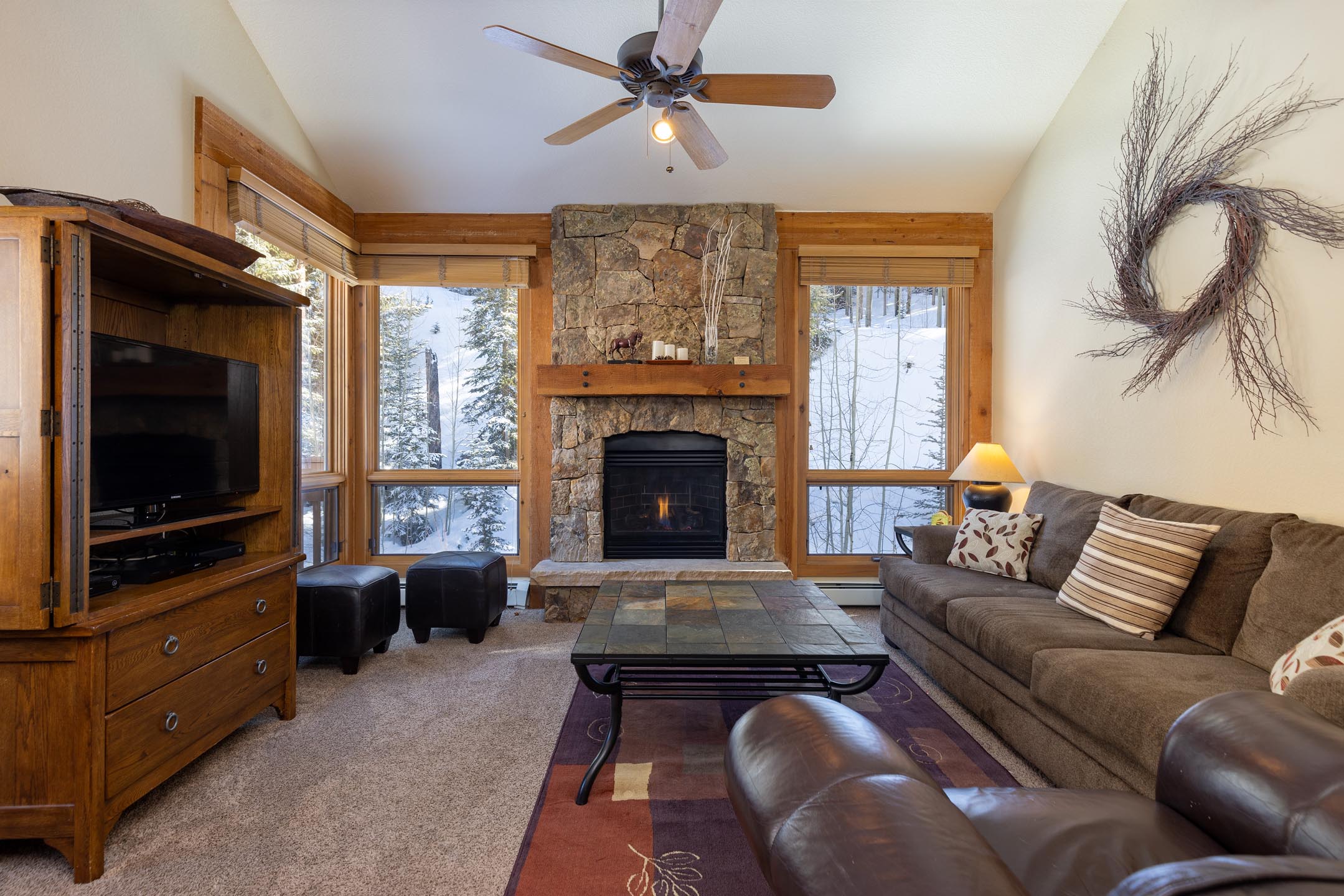 A cozy living room with a stone fireplace, a wall-mounted TV, wooden furniture, a brown sofa, and a large window showing a snowy landscape outside.