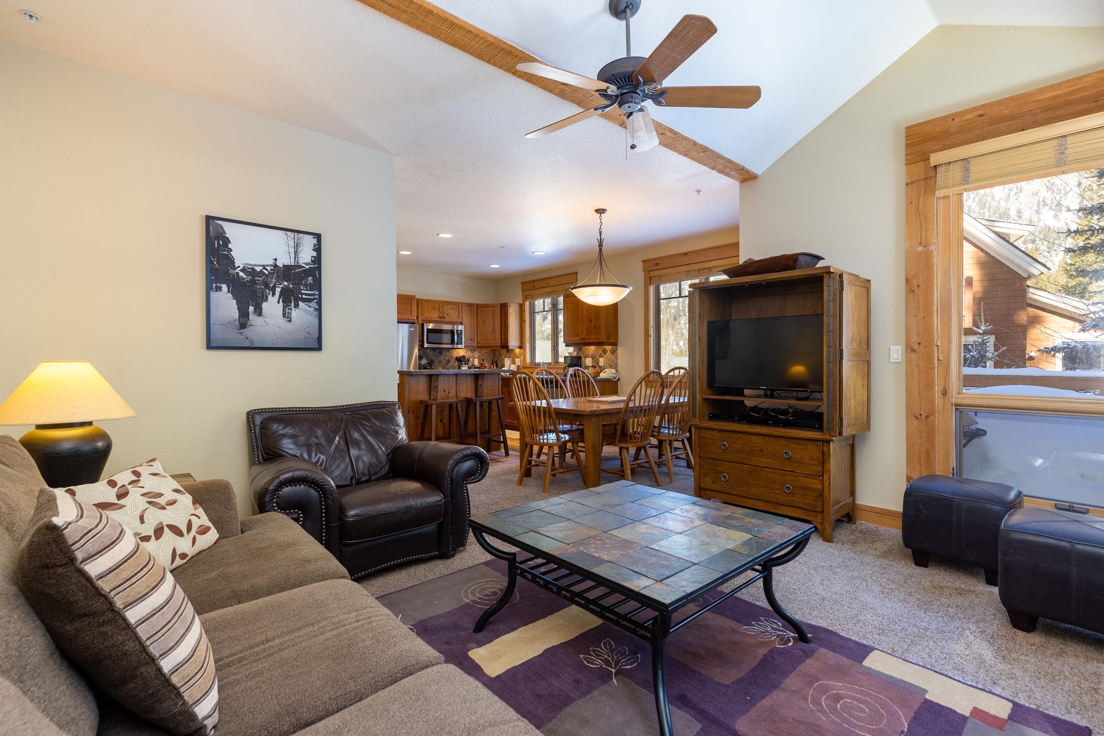 Living room with a leather armchair, cushioned sofa, coffee table, and TV. Dining area with wooden table and chairs visible in the background. Ceiling fan and bright natural light from windows.