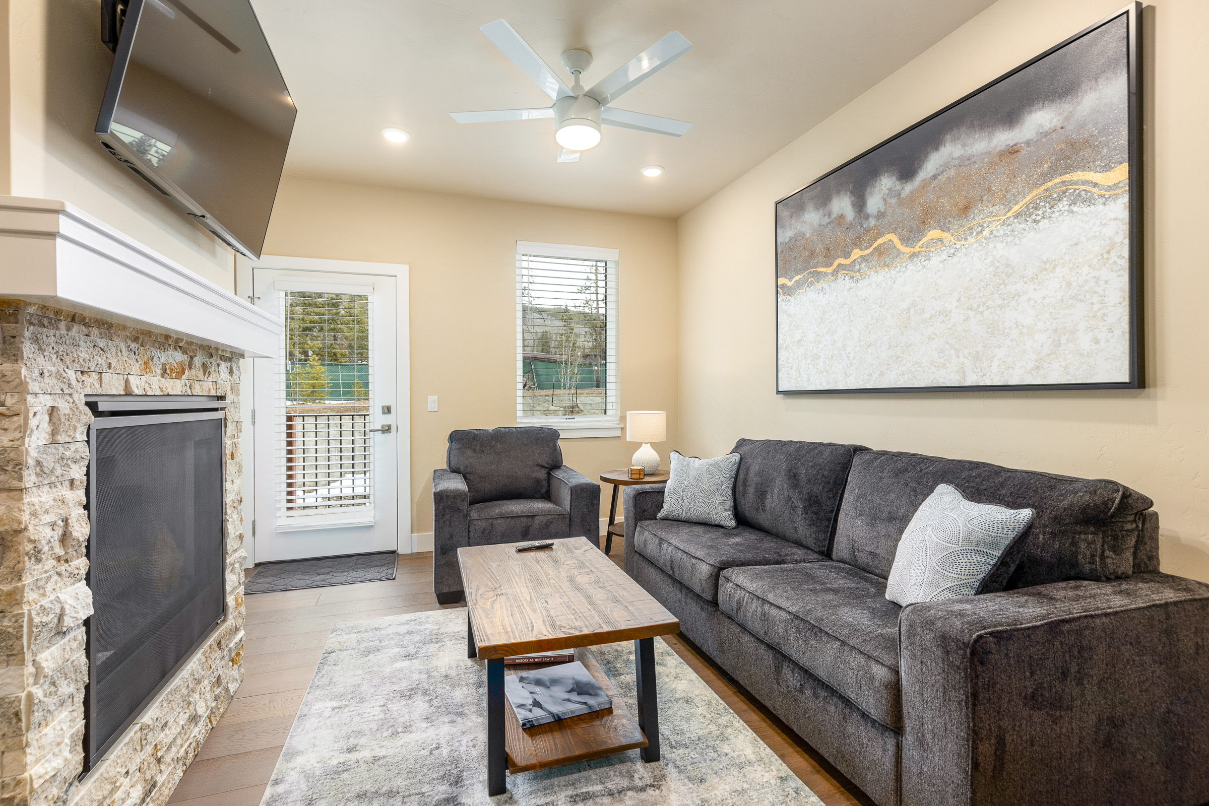 A living room with a gray sofa, two armchairs, a wooden coffee table, and a TV mounted above a stone fireplace. Artwork hangs on a beige wall, and a door with a window leads outside.