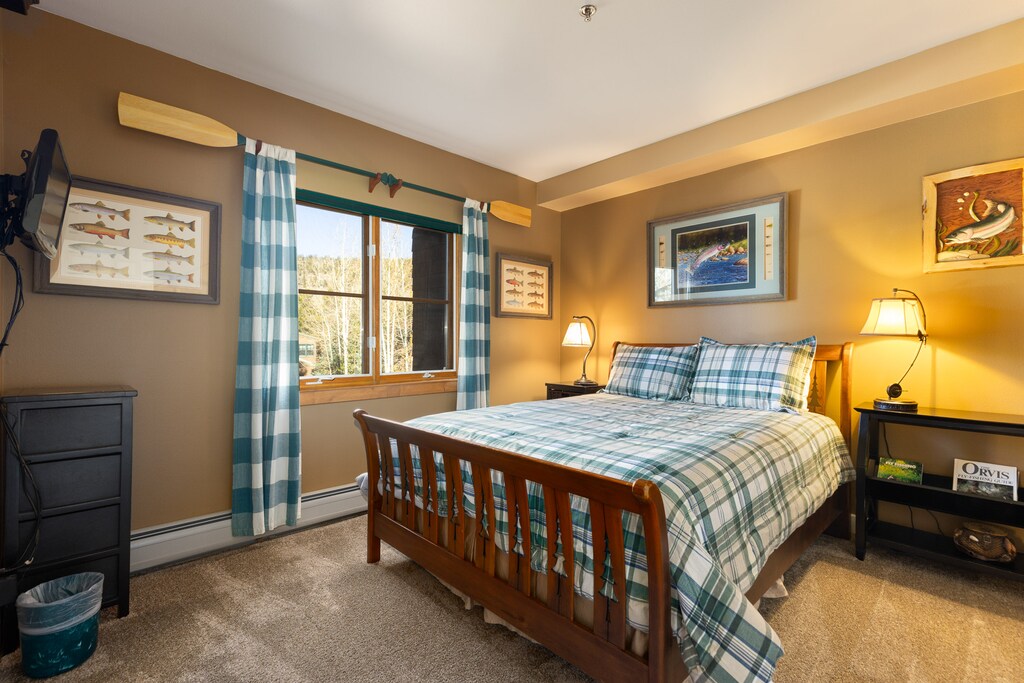 A bedroom with a wooden bed frame, plaid bedding, two bedside tables with lamps, checkered curtains, and fishing-themed wall decorations. There is a wall-mounted TV and a window with a scenic view.