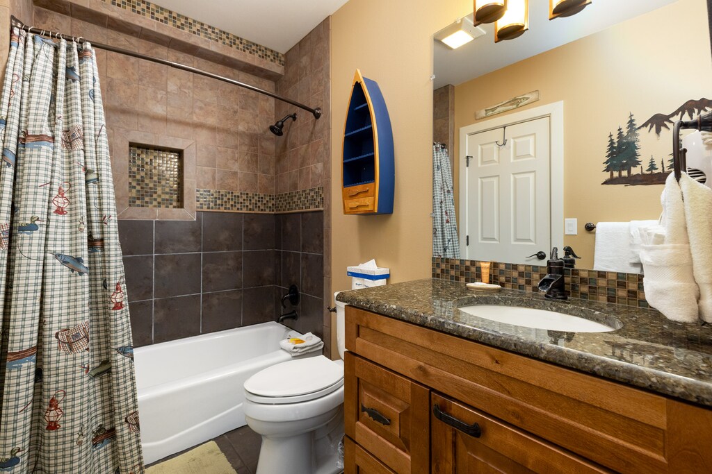 A bathroom with a tub-shower combination, a vanity with granite countertop, a wall-mounted boat-shaped shelf, and a nature-themed shower curtain.
