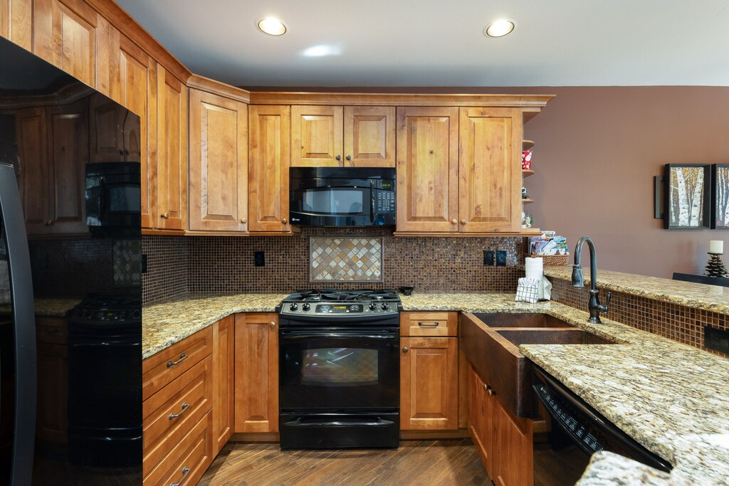 Modern kitchen with wooden cabinets, granite countertops, black appliances, and a tile backsplash. There is a built-in microwave above the stove and a double sink.
