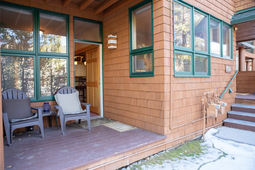 A wooden cabin porch with two chairs and small table, glass windows, a partially open door, and a few steps leading up to the entrance. There is some snow on the ground near the porch.
