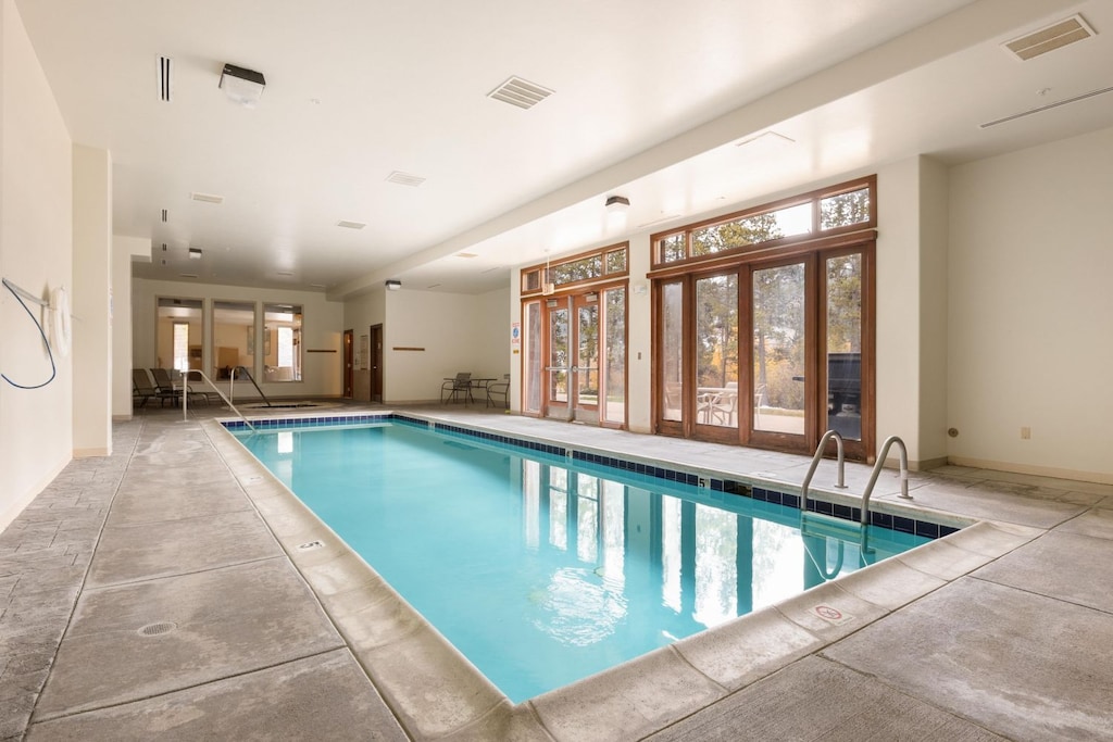 Indoor swimming pool with concrete decking and lounge chairs along one side. Large windows and glass doors on one wall provide natural light and a view of an outdoor area.