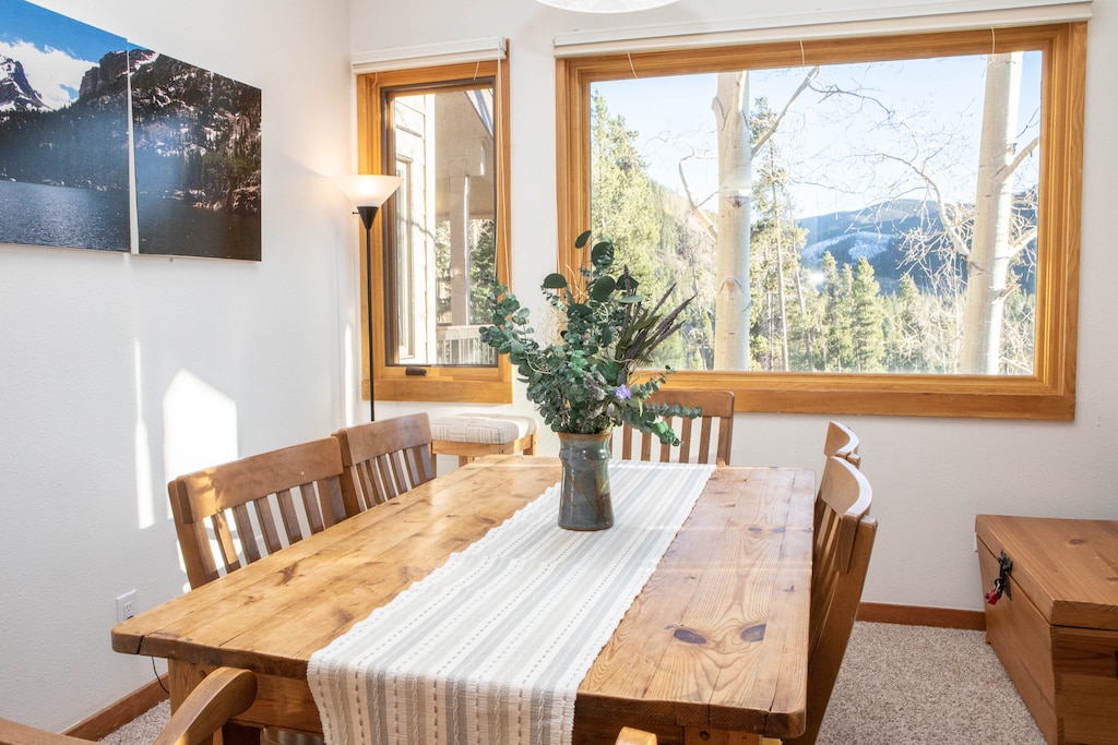 A dining room with a wooden table and chairs, a vase with greenery as a centerpiece, and a large window offering a mountain view. A framed photo is visible on the wall, and a floor lamp stands in the corner.