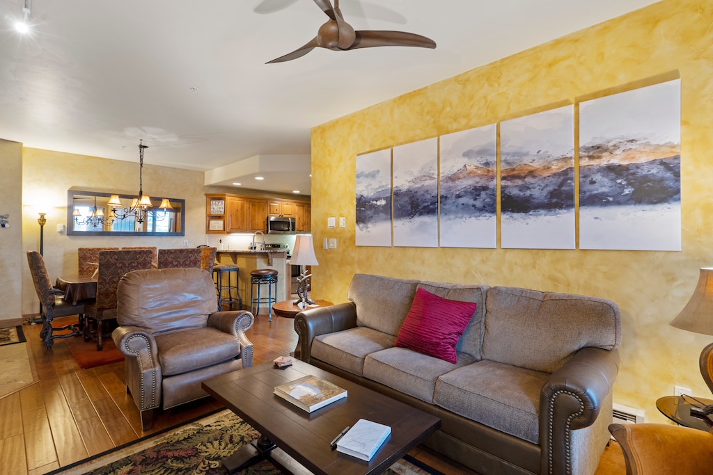 Living room with a brown sofa, two armchairs, a wooden coffee table, and a multi-panel painting on a yellow wall. A kitchen and dining area are visible in the background.