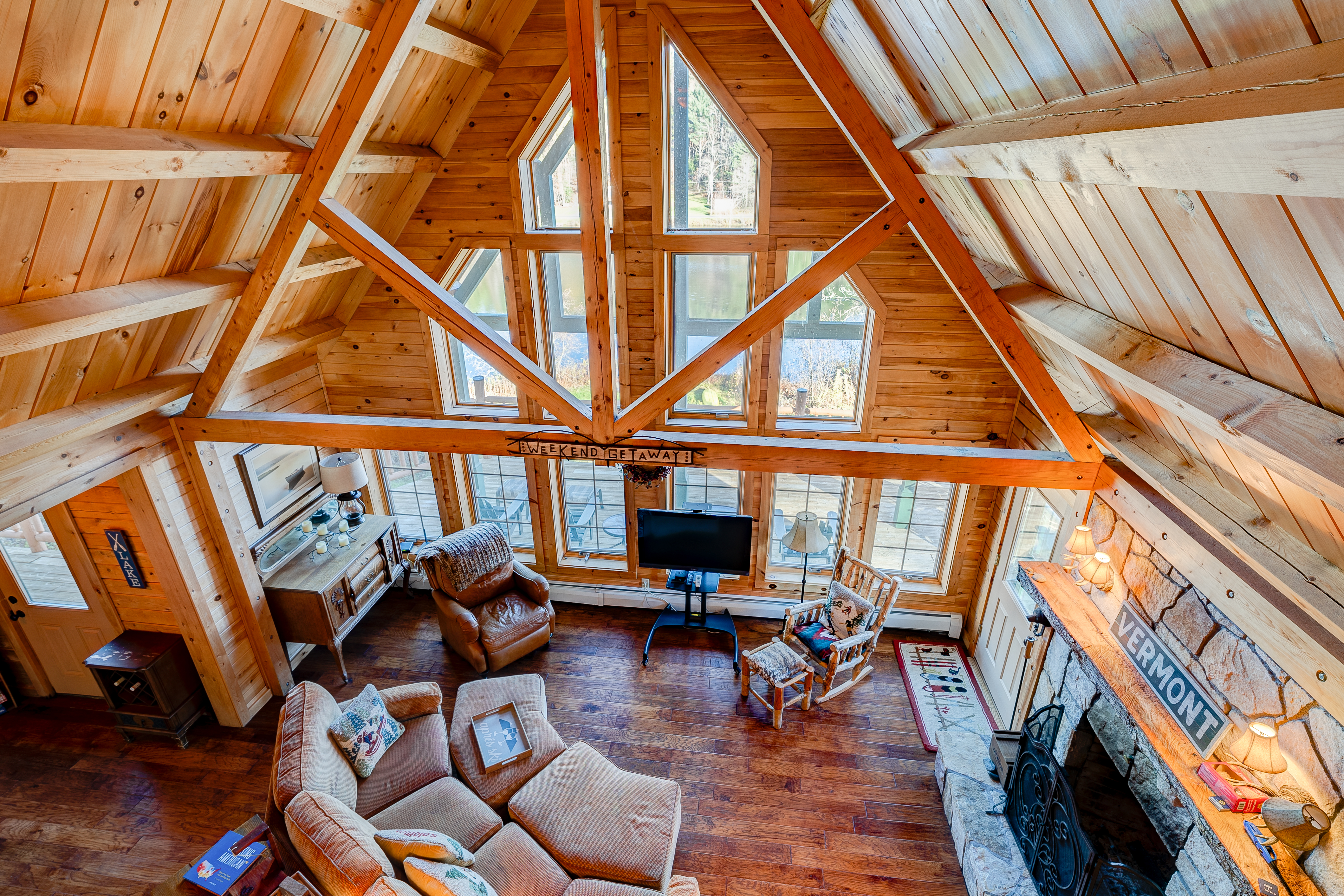 A cozy wooden cabin living room with vaulted ceilings, large windows, a TV, a fireplace, and various seating options including armchairs and a sofa.