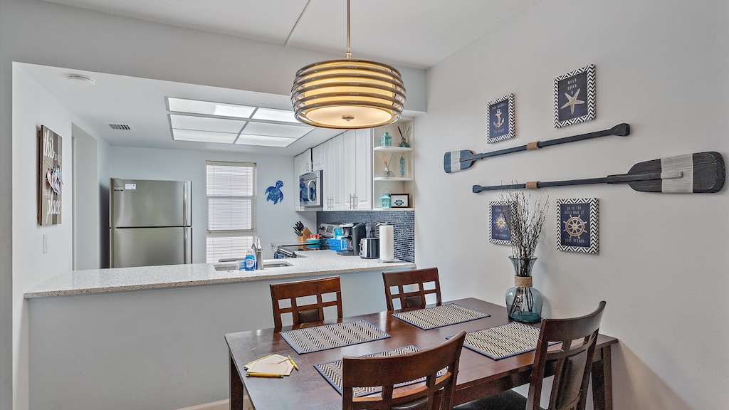 A small, modern kitchen and dining area with a wood table, four chairs, wall decor of paddles, and nautical-themed art. The kitchen features a stainless steel refrigerator and an open shelf design.