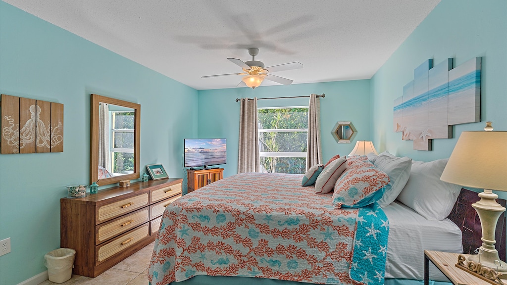 A bedroom with turquoise walls, wood furniture, and a bed with coral-themed bedding. The room has a ceiling fan, a TV on a dresser, a window with gray curtains, and beach-themed decor.