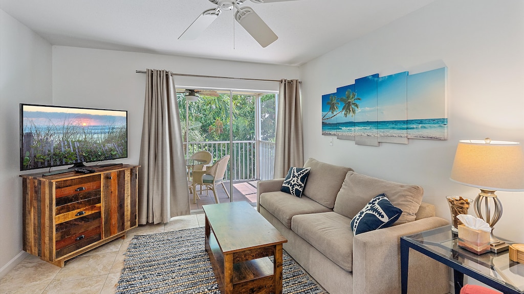 A cozy living room with a gray sofa, blue cushions, a wooden coffee table, a TV on a wooden stand, a ceiling fan, beach-themed artwork, and a sliding door leading to a balcony with outdoor seating.