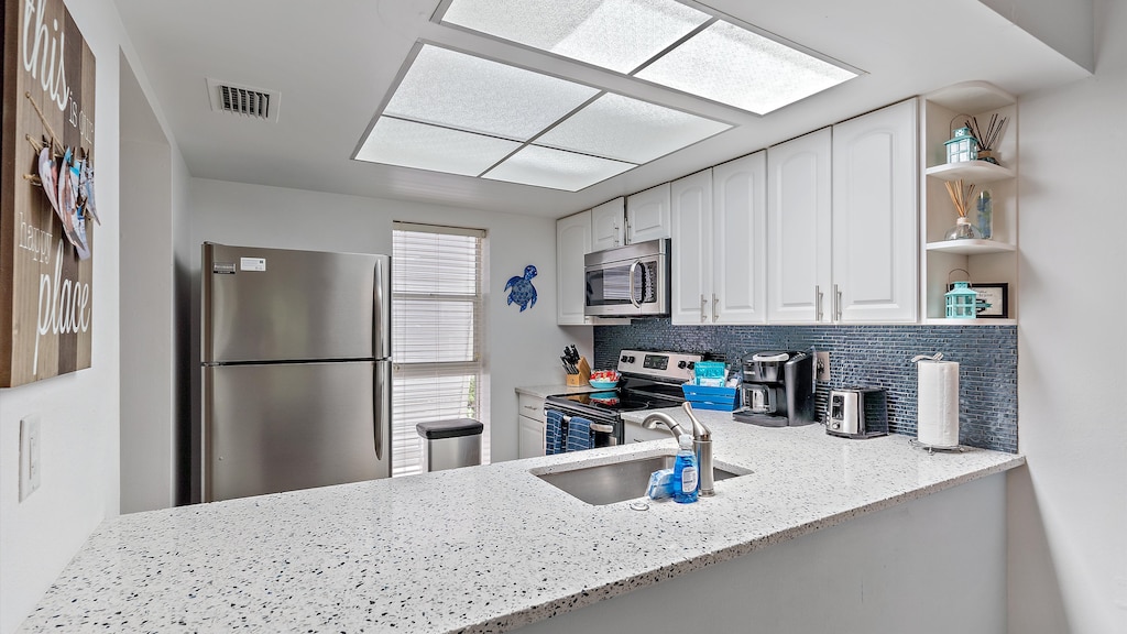 A modern kitchen featuring a stainless steel refrigerator, stove, and microwave, blue tiled backsplash, white cabinets, and a speckled countertop with a sink.