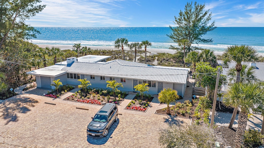 A single-story beachfront house with a gray roof and a landscaped front yard, featuring a single SUV parked in the driveway. The ocean is visible in the background under a blue sky.