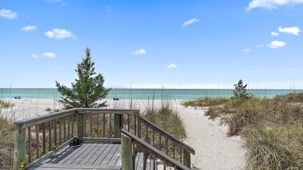 Wooden pathway leading to a sandy beach with a clear blue sky and calm ocean in the background. Sparse vegetation lines the path, and a pair of sandals is placed on the wooden boards.