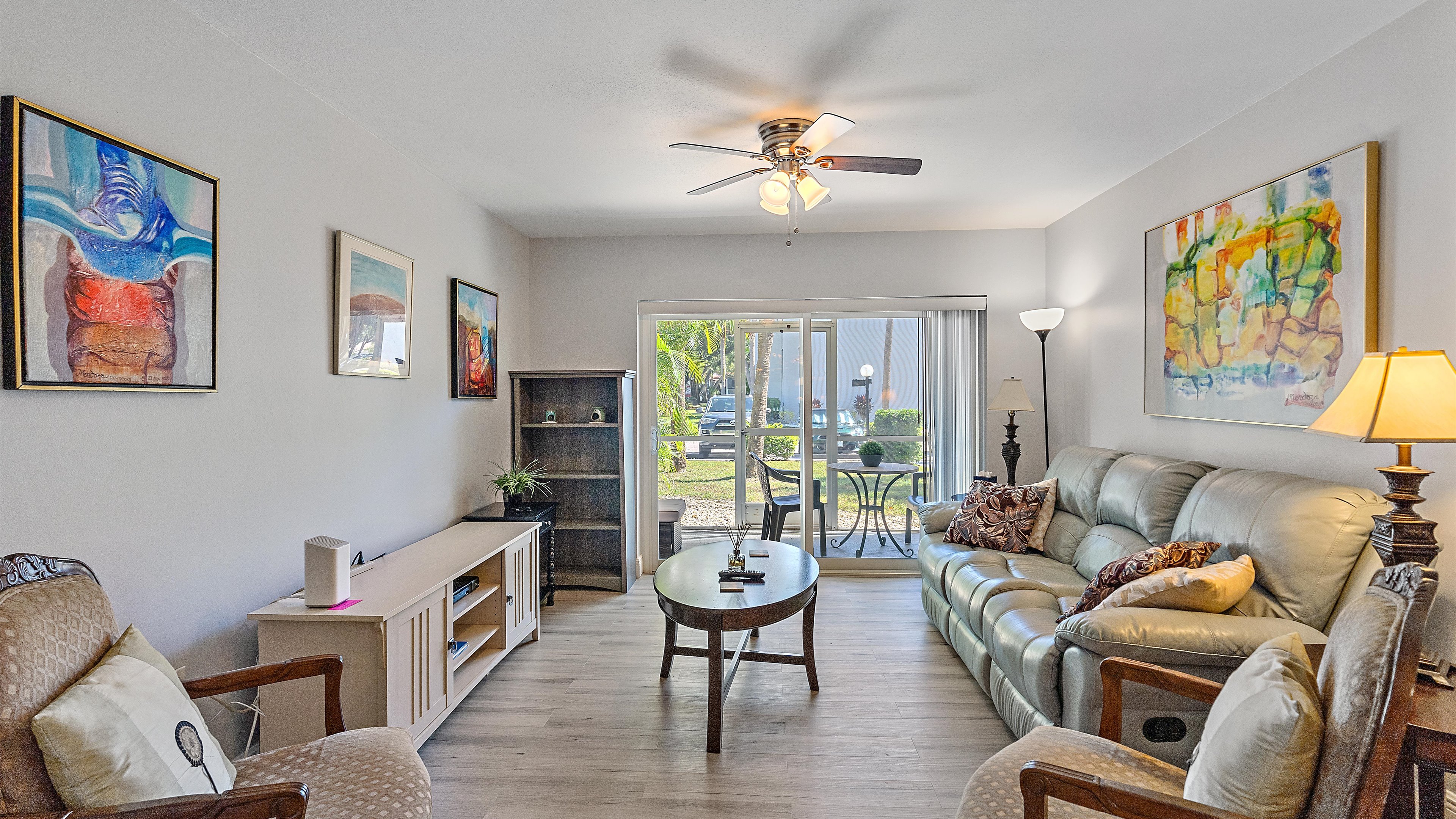 A cozy living room with a ceiling fan and art on the walls. It has a sofa, chairs, a wooden coffee table, an entertainment unit, and a glass door leading to a patio with a garden view.