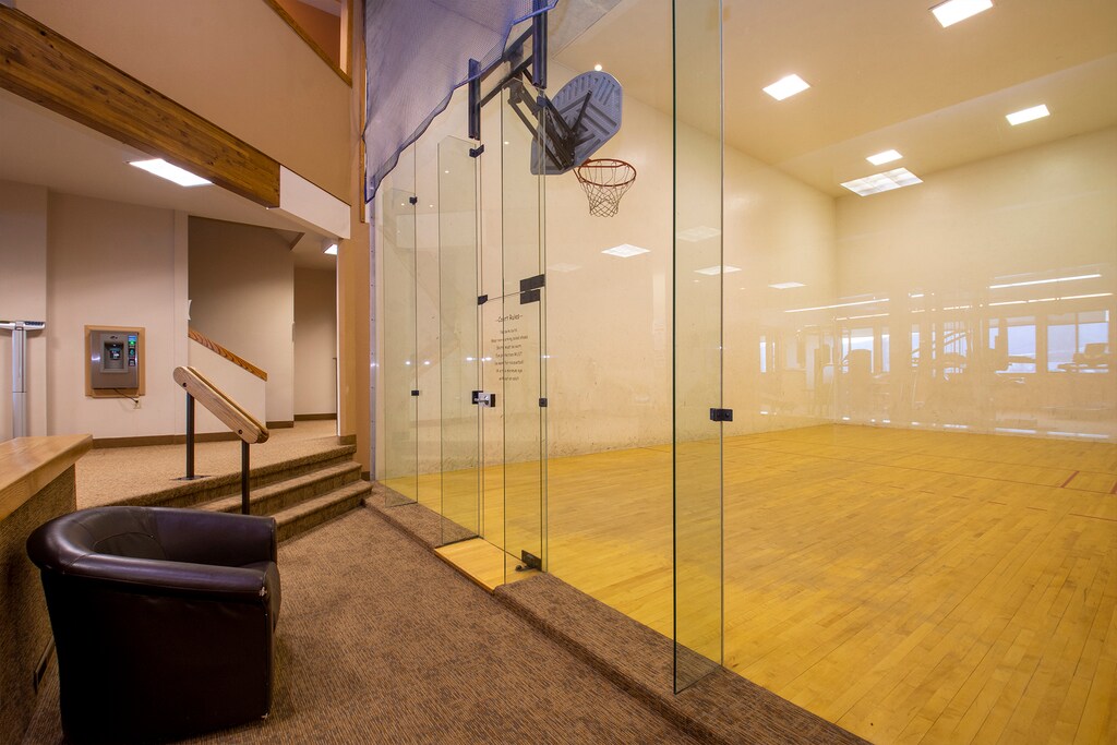 A clean, indoor squash court with glass walls, wooden flooring, and a small basketball hoop on one wall. A black leather chair is placed outside the court near the entrance.