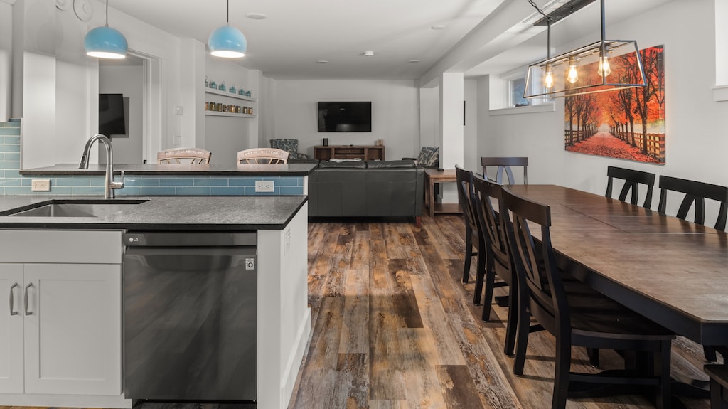 An open-concept kitchen and dining area with a sink and dishwasher on the left, a long dining table with chairs on the right, and a seating area with a sofa and TV in the background.
