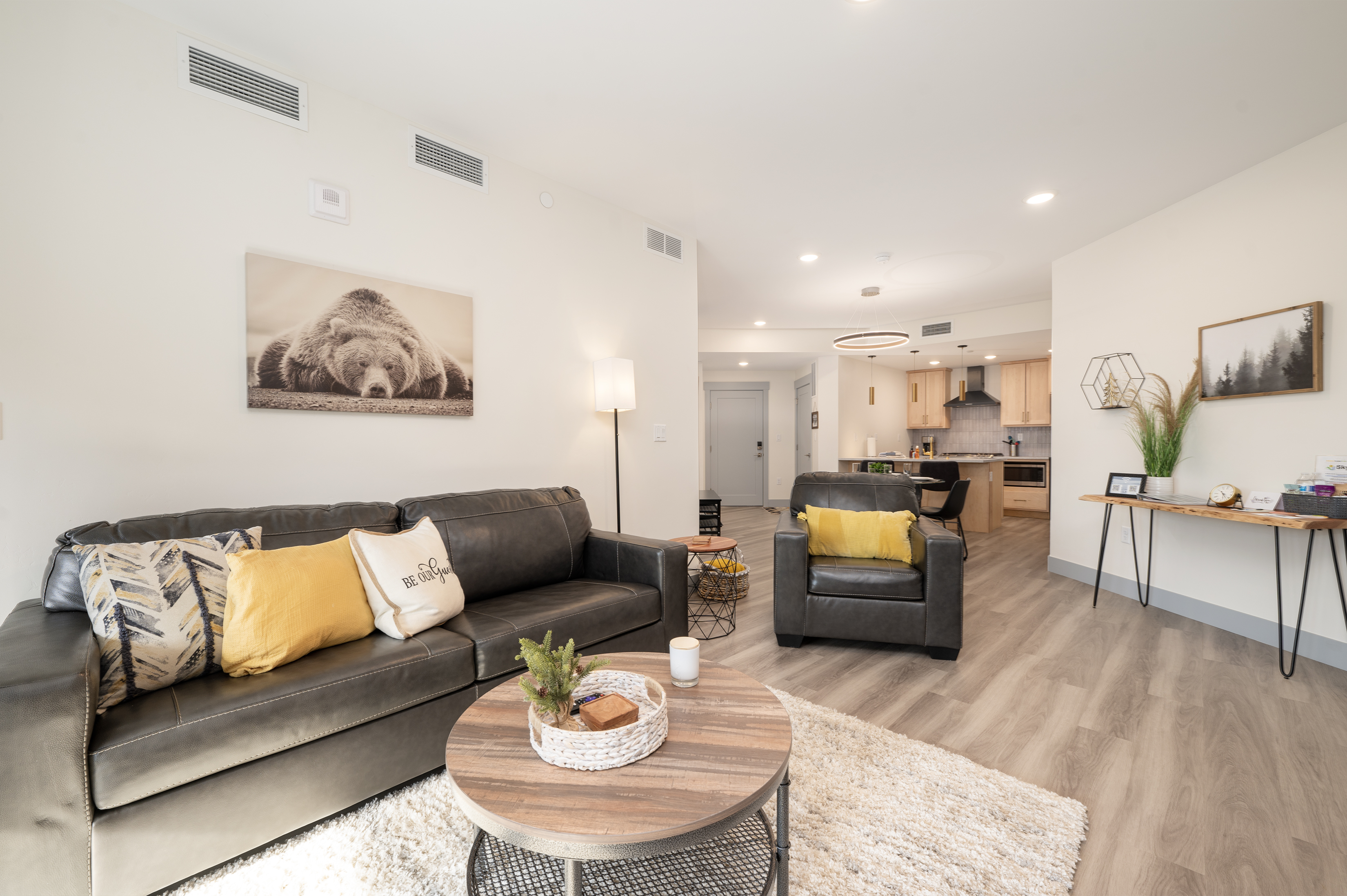 A modern living room with a black couch, armchair, and wooden coffee table. The room features plants, wall art, yellow cushions, and an open view of the kitchen area in the background.