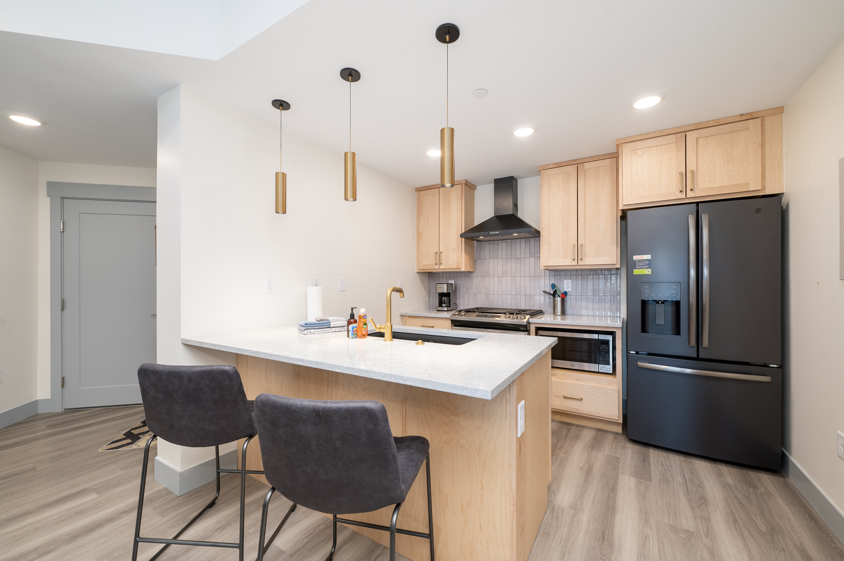 A modern brand new kitchen featuring light wood cabinets, a black refrigerator, a black oven range with a hood, a grey backsplash, and a white island with two grey barstools.