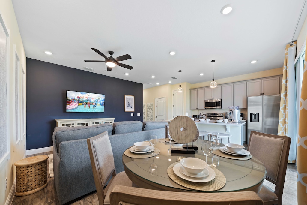 Cozy open-concept dining area with navy accent wall and mounted TV - Dining table set for four, pendant lights over a modern kitchen with stainless steel appliances - Natural light from large windows with patterned curtains brightens the space
