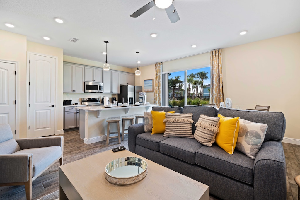 Stylish living area featuring a modern kitchen with stainless steel appliances and a breakfast bar with stools - Comfortable gray sofa adorned with yellow and patterned pillows - Large sliding glass door revealing a scenic view of sunny outdoors