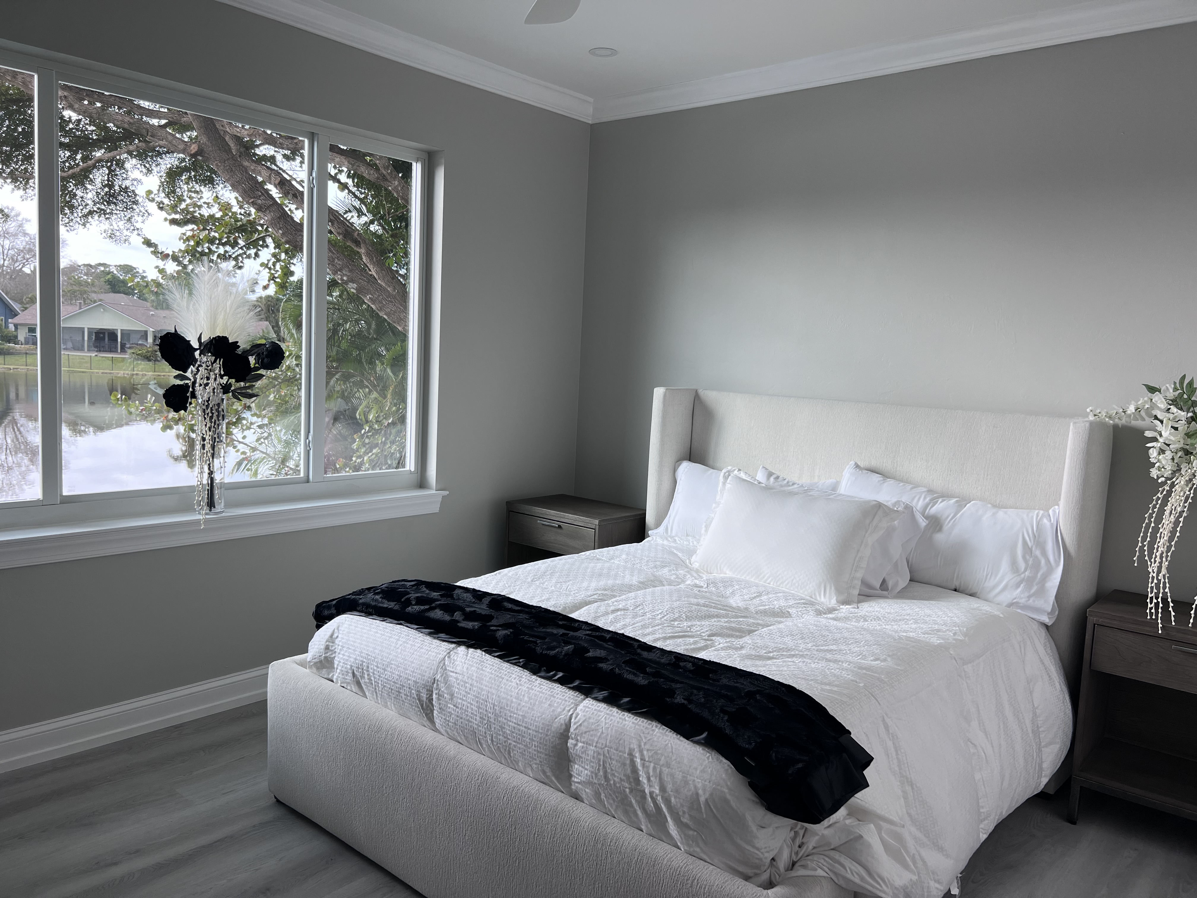 A bedroom with a queen-sized bed, white bedding, black decorative pillow, and two wooden nightstands near a large window overlooking a lake and trees.