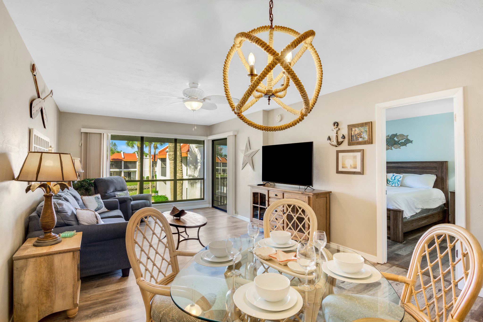 A dining area with a round table set for four, adjacent to a living room with a sofa, TV, and sliding glass doors leading to a balcony. A bedroom is visible through an open doorway on the right.