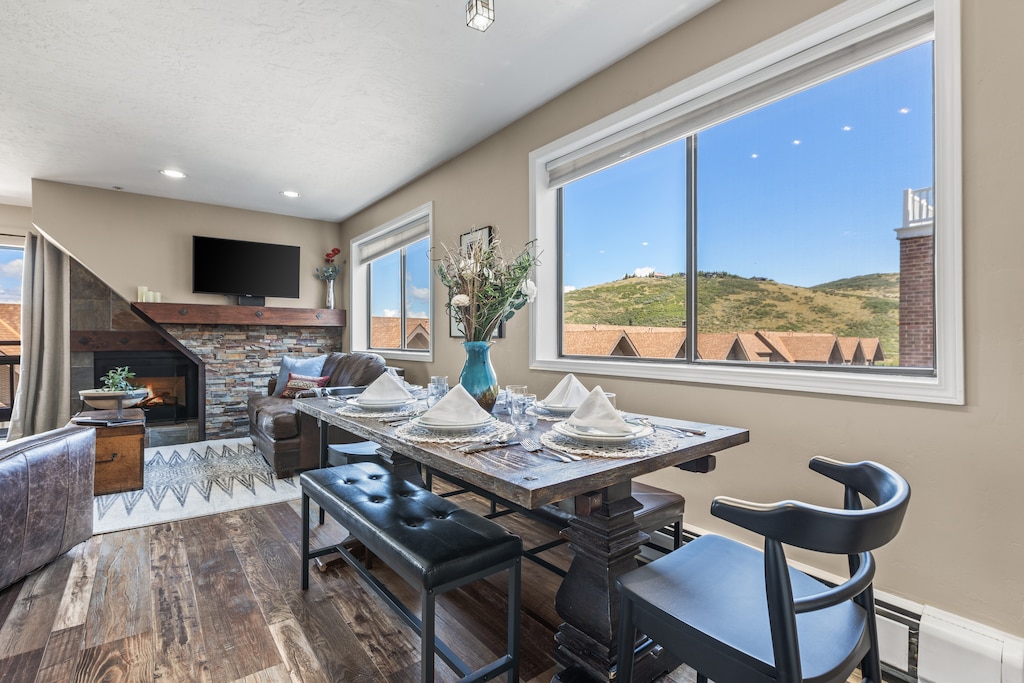 A dining area with a wooden table set for four, adjacent to a living room with a leather sofa, stone fireplace, and large windows showing a scenic mountain view.