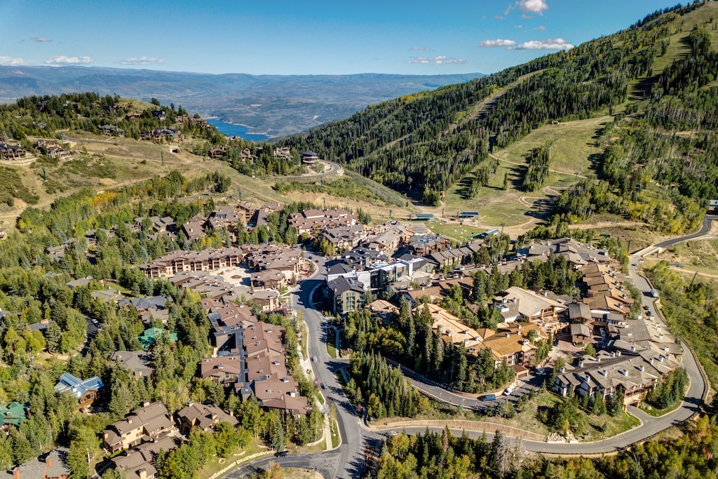 Mountain resort village nestled in a scenic valley with ski slopes, surrounded by forested hills and distant peaks under blue skies.