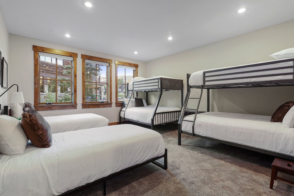 Bedroom with two sets of bunk beds and two single beds, large windows with wooden frames, and neutral-colored walls and carpet.