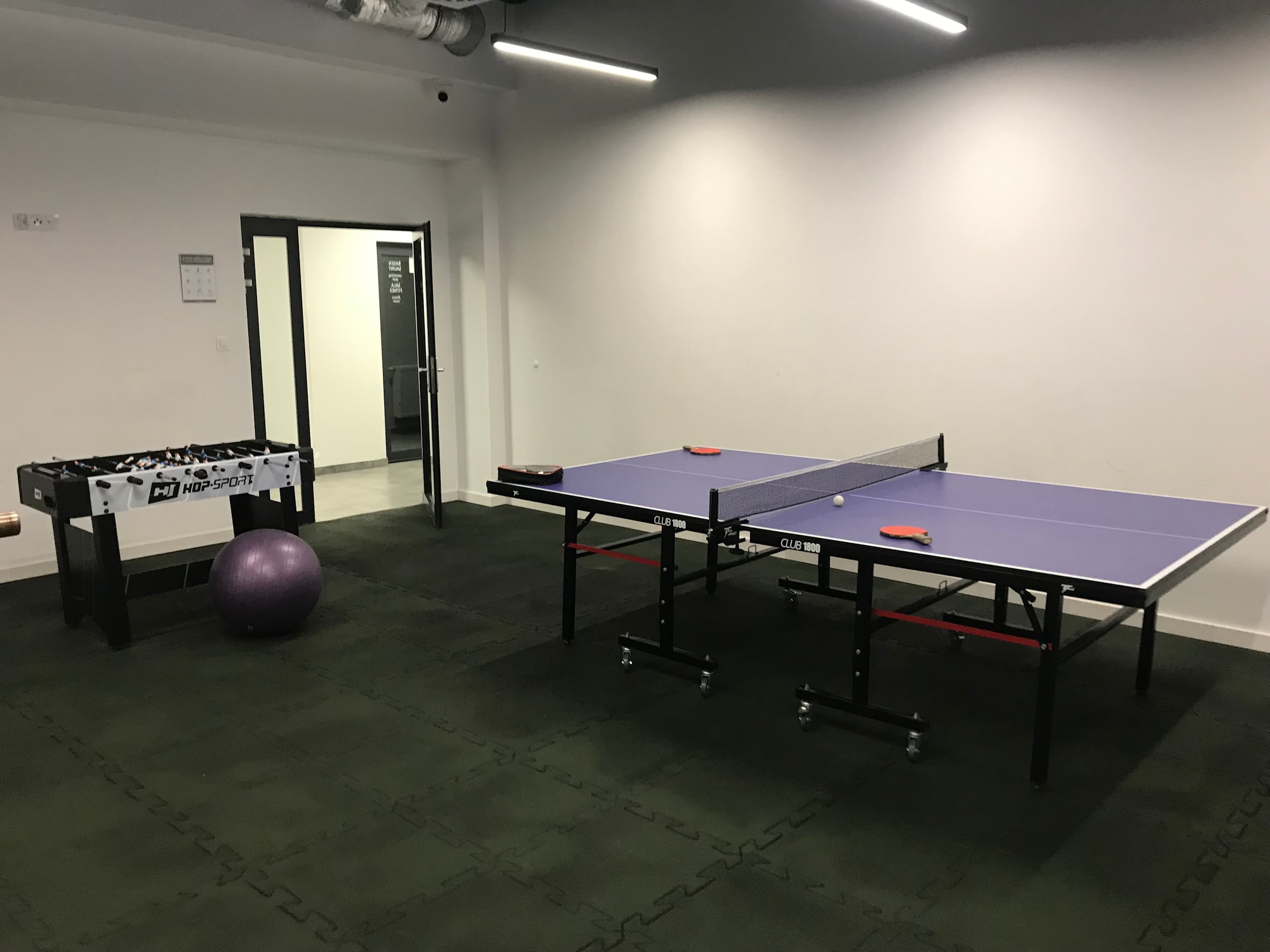 Multifunctional fitness room combining gym equipment, cardio machines, and free weights with recreational elements like table tennis and foosball. 