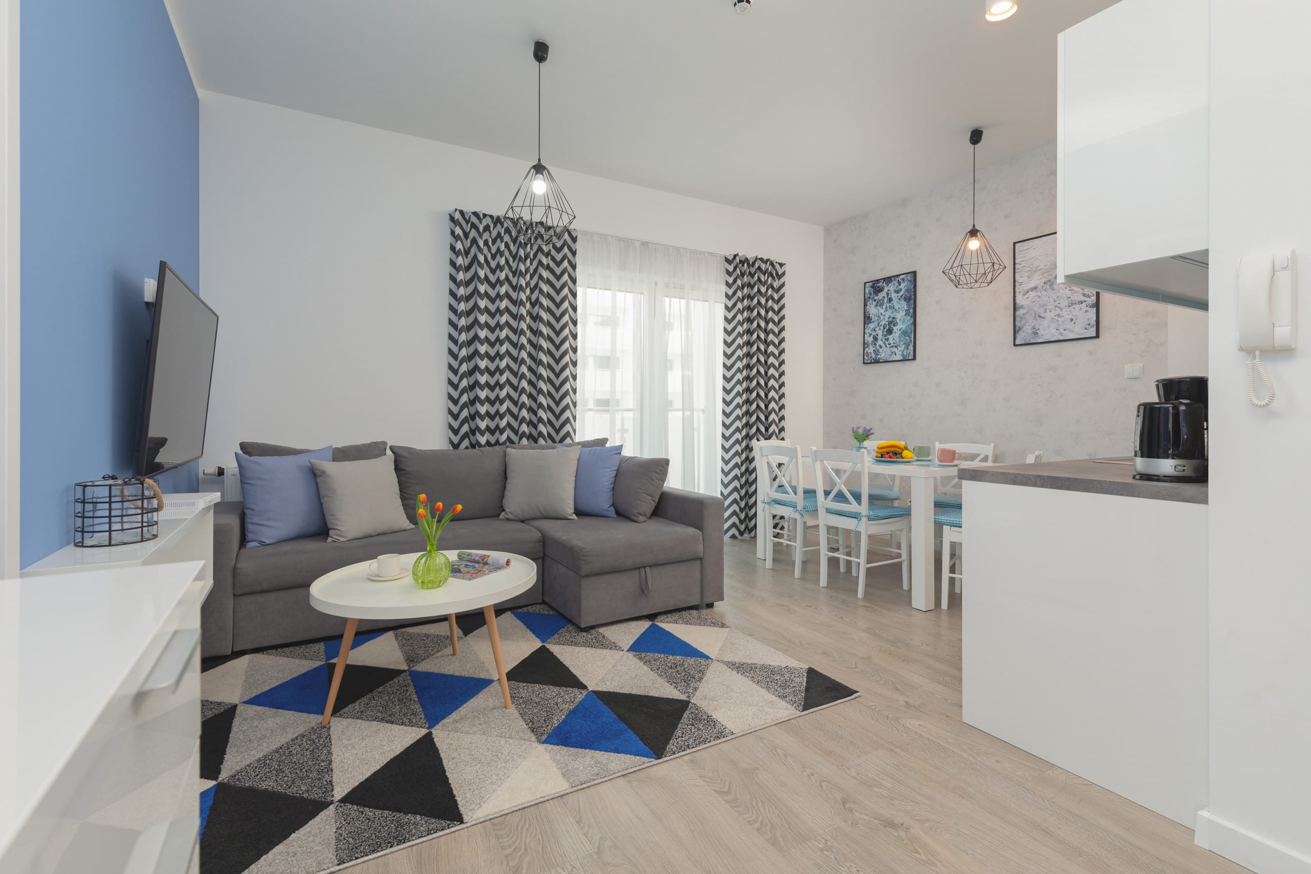 A cozy living room with a blue and gray theme, featuring a large sofa, a stylish rug, and a mounted TV. The decor gives a modern and welcoming feel.