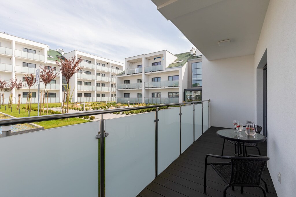 A balcony view overlooking a serene residential area with white modern buildings and greenery. The balcony has simple yet functional seating.