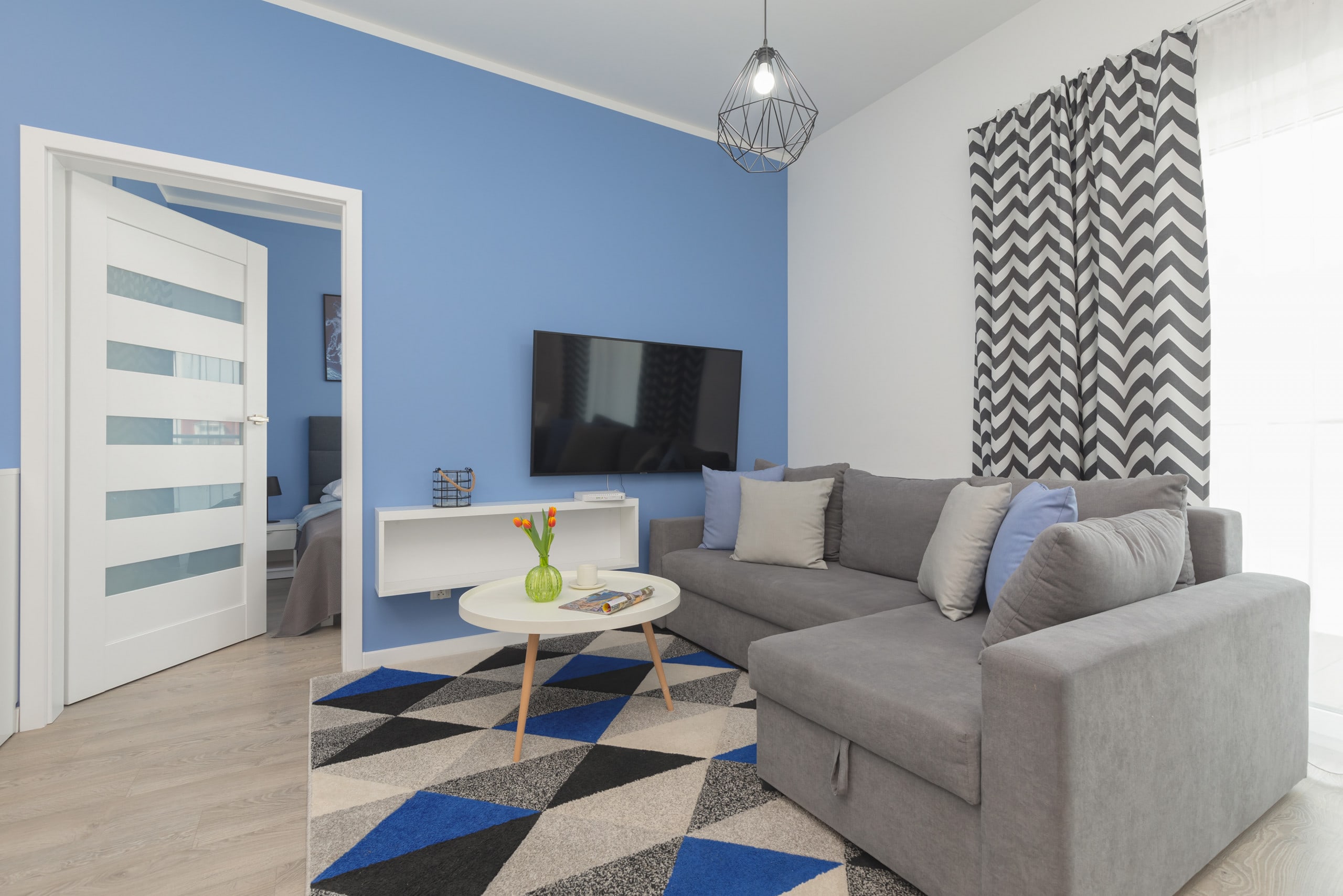 A bright and spacious living room featuring a comfortable gray sofa, a stylish patterned rug, and a mounted TV. Large windows fill the space with natural light, enhancing the modern yet cozy ambiance.