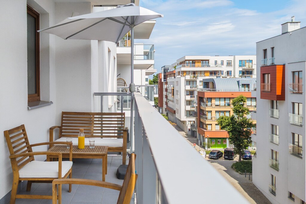 A serene balcony space with wooden chairs and a table, overlooking a colorful modern residential area. Perfect for relaxing outdoors with a coffee or book.