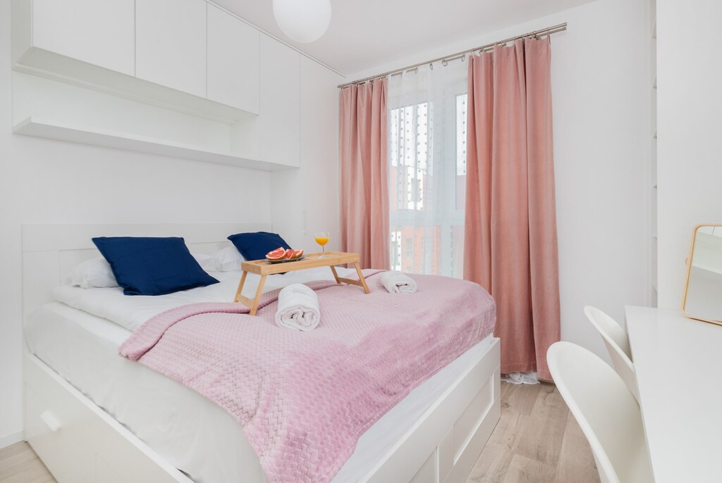 A charming bedroom featuring a soft pink bedspread and neatly arranged pillows, lit by natural light through large windows. Curtains in soft pink tones add to the inviting ambiance.