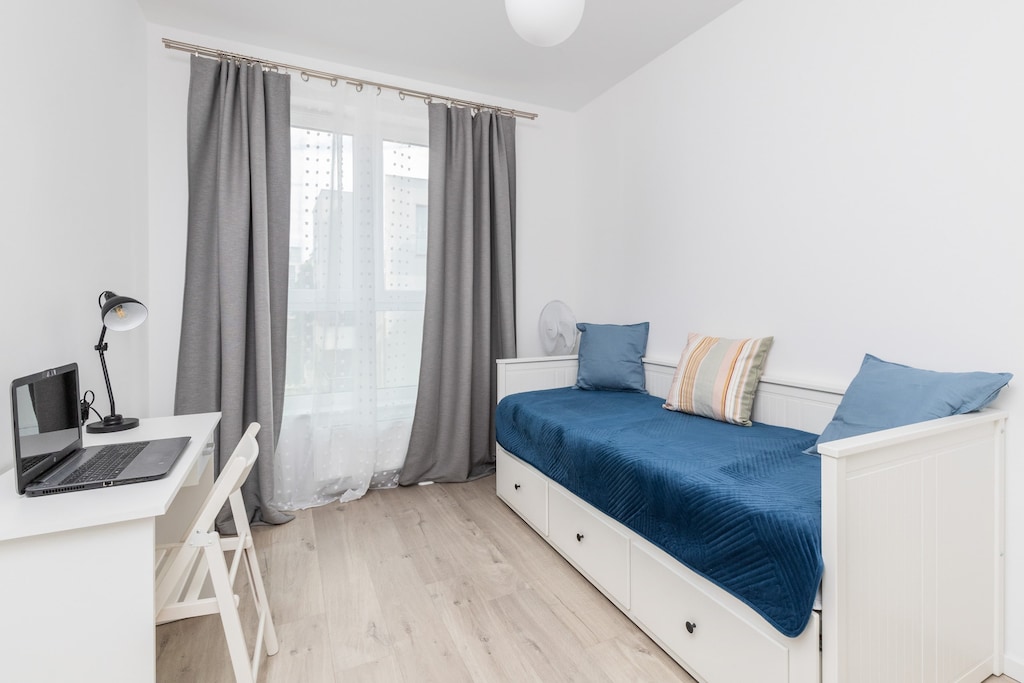 A bright single-bedroom setup with a neatly made blue bed and minimalist furniture. The window and curtains allow ample daylight to fill the space.