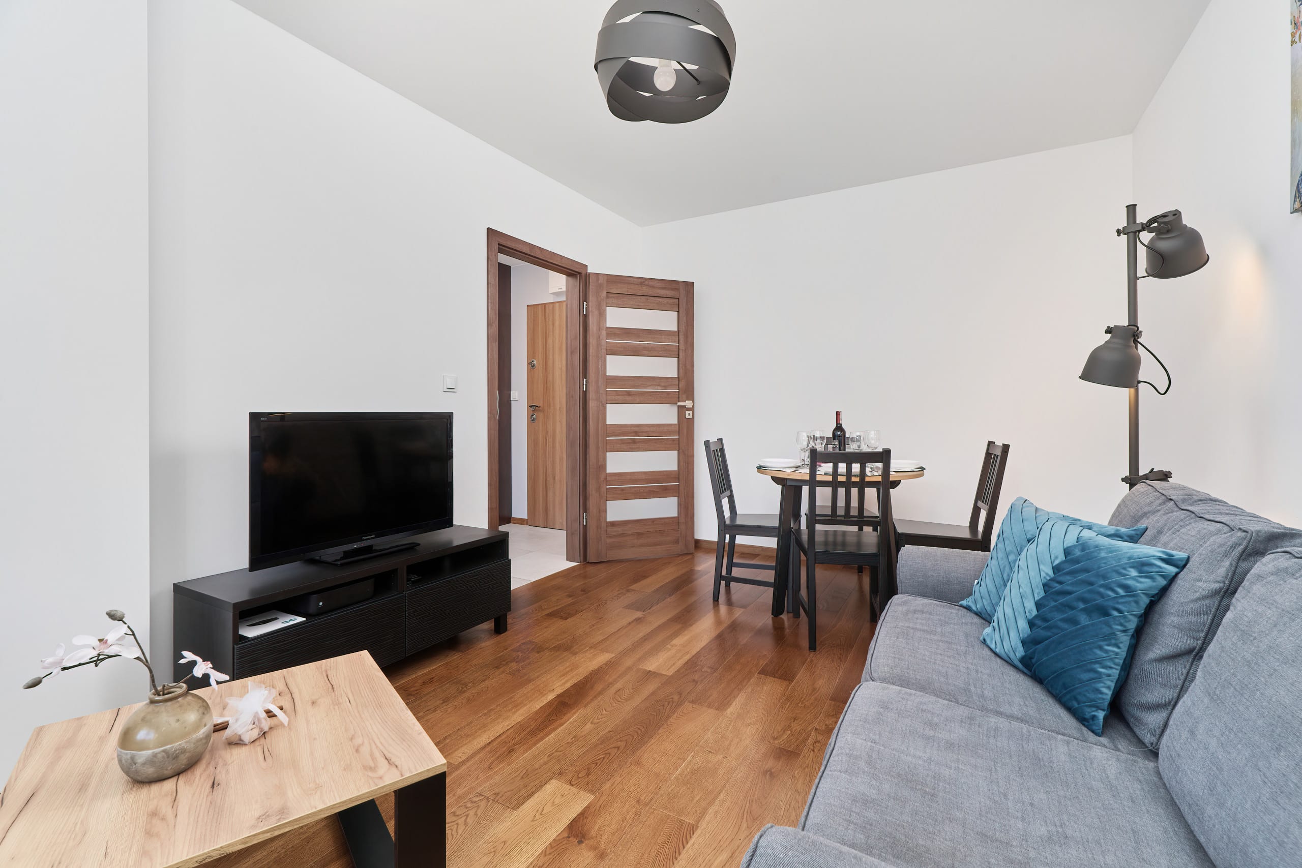 A spacious dining and living area, with a wooden table set for four, a gray sofa, and wall art adding personality to the room.