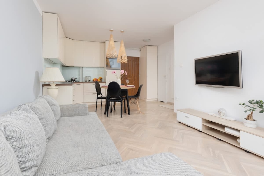 A spacious living room with a large gray sofa, a flat-screen TV mounted on the wall, and natural light filling the space from the windows, creating a relaxing ambiance.