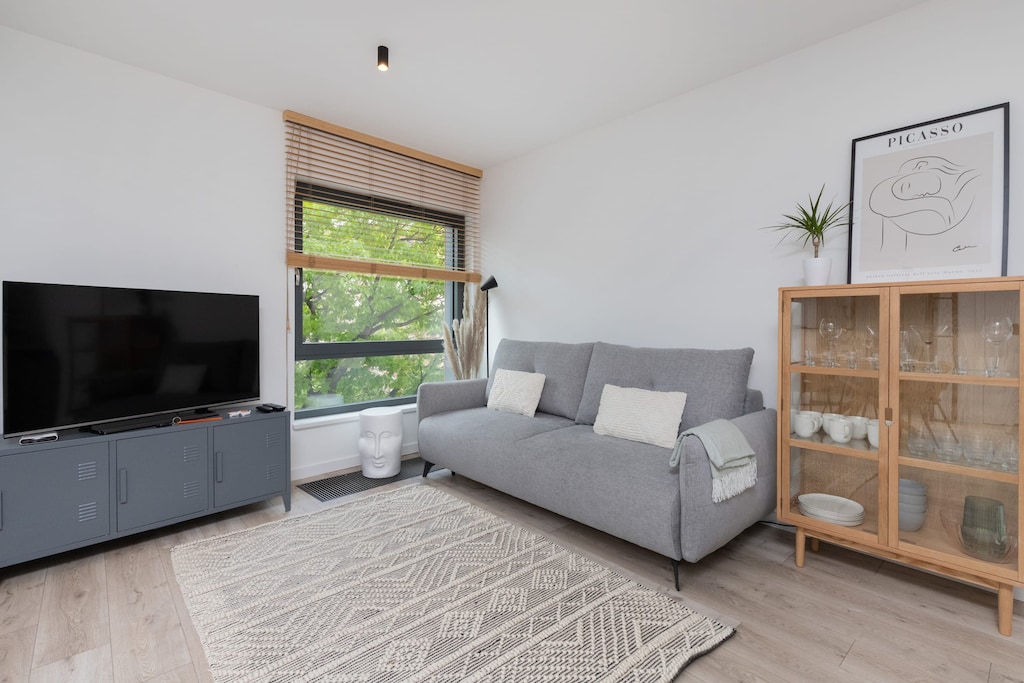 A cozy living room with a gray sofa, a wooden side table, and a minimalistic bookshelf, complemented by soft natural light from large windows.