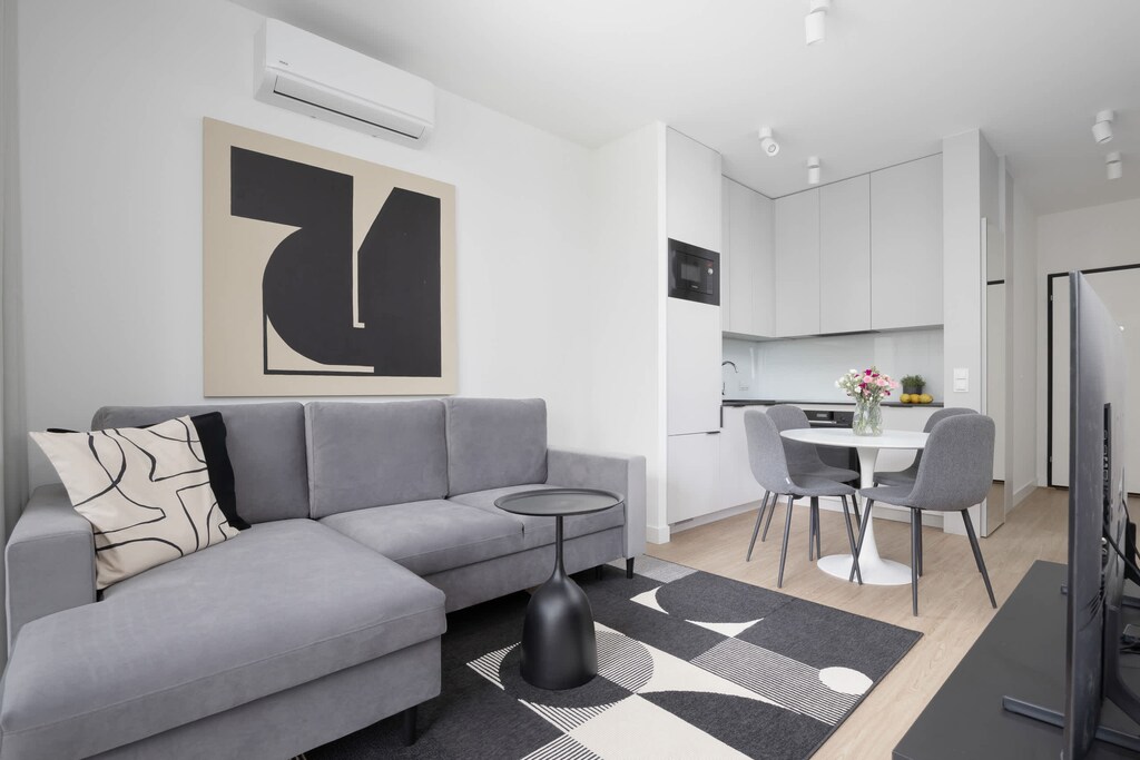 A bright living room featuring a sectional sofa, small dining table, and white interior palette with minimalist decor.